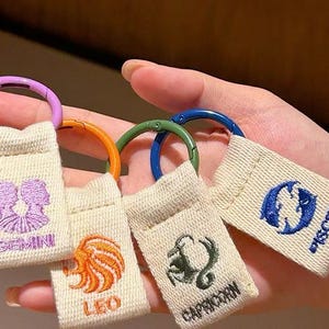 Puede incluir: Cuatro llaveros rectangulares de tela color crema con signos del zodiaco y texto bordados. Cada llavero tiene una anilla de metal de color: morado para Géminis, naranja para Leo, verde para Capricornio y azul para Piscis.