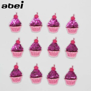 May include: Twelve cupcake-shaped patches are displayed. Each patch has a pink cupcake case, purple sequined frosting, and a red cherry. The patches are outlined in white and set against a white backdrop. The word "abei" is in the top left corner.