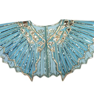 May include: A light blue sheer capelet featuring a scalloped hem and intricate gold sequin detailing arranged in radiating lines and geometric patterns across the fabric. The garment has a wide, open neckline and a structured, pleated design.