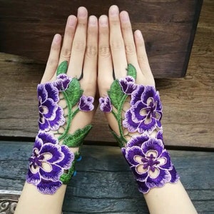 Puede incluir: Un par de puños de encaje floral morado. Los puños presentan flores moradas bordadas con hojas y tallos verdes. El diseño calado crea un aspecto delicado y elegante.