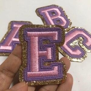 May include: Embroidered letter patches in pink and purple with gold glitter borders. The letter "E" is in focus, with "A", "B", and "C" visible in the background. These patches are suitable for various craft projects.