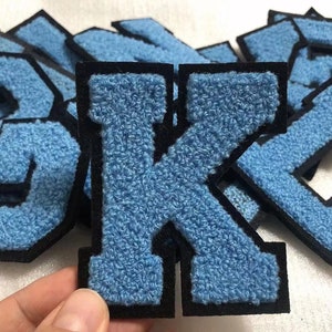 Könnte beinhalten: Nahaufnahme eines blauen Filzbuchstabens K mit schwarzen Nähten. Der Buchstabe ist von anderen Buchstaben umgeben, die alle aus blauem Filz mit schwarzen Nähten bestehen.