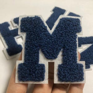 Può includere: Quattro toppe di lettere in feltro blu navy con contorni bianchi. Le lettere sono H, M, Z e una M più grande.