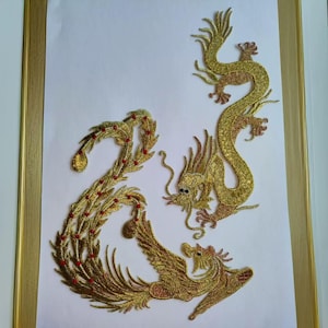 May include: A framed artwork featuring gold embroidered mythical creatures. A dragon and a phoenix are depicted in intricate detail with red accents. The artwork is set against a white background and framed in a gold-colored frame.
