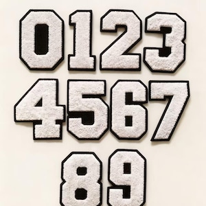 Puede incluir: Un conjunto de parches de números blancos y esponjosos, estilo universitario, que van del 0 al 9. Cada número tiene un borde negro y una apariencia texturizada y difusa. Los números se muestran sobre un fondo blanco liso.