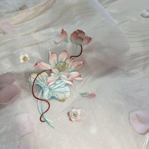 Op de afbeelding: Een doorschijnende witte stof met delicaat borduurwerk van een lotusbloem in zachte roze, blauwgroene en gouden draden. Het ontwerp bevat een centrale lotusbloem, een groot blad en kleinere bloemaccenten, aangevuld met verspreide lichtroze bloemblaadjes.
