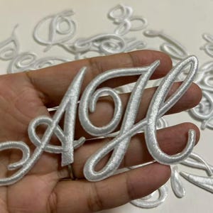 May include: White embroidered letters, including "A" and "H", are displayed against a light background. The letters are in a cursive script style, with a satin-like texture. Other letters are scattered around, suggesting a collection of alphabet patches.