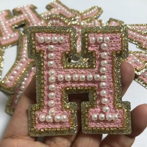 May include: Pink felt letter H with a gold glitter outline and white pearl embellishments. The letter is surrounded by other letters in the same style.