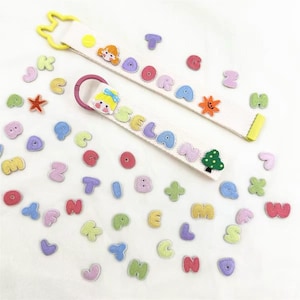 Puede incluir: Dos correas de tela blanca con clips de plástico, uno amarillo y otro rosa. Las correas presentan parches de letras coloridas y texturizadas que forman ADORA y SELAN, junto con pequeños iconos como un sol y un árbol, rodeados de letras sueltas.