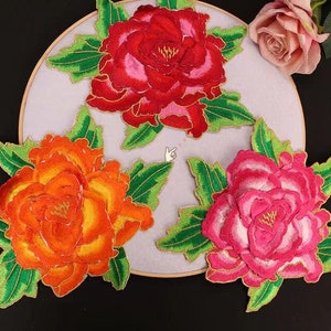 Puede incluir: Tres aplicaciones de flores bordadas en tela en rosa, naranja y rojo con hojas verdes. Las flores son de estilo peonía.