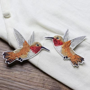 May include: Two embroidered iron-on patches of hummingbirds in flight. The hummingbirds are orange, red, and white with gray wings.