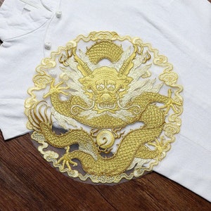 May include: A gold embroidered dragon with white accents on a white mesh background. The dragon is curled up in a circle with its head facing forward. The dragon is surrounded by clouds.