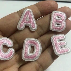 Puede incluir: Letras de tela bordadas A, B, C, D y E en color rosa suave con borde blanco. Estos parches termoadhesivos son ideales para personalizar ropa, bolsos u otros artículos de tela. Las letras están en una fuente redondeada sin serifa.