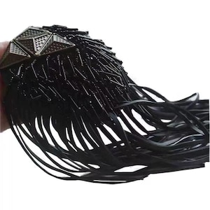 Peut inclure: Un collier noir en perles avec un pendentif géométrique et une longue frange. Le pendentif est en métal et a une finition argentée.