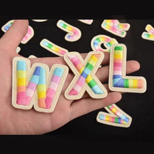 Puede incluir: Parches bordados con las letras W, X y L en un esquema de colores arcoíris. Los parches tienen un borde color crema y están colocados sobre un fondo oscuro. También se ven otros parches de colores arcoíris.