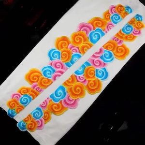 Puede incluir: Dos tiras de tela blanca con diseños de nubes bordadas de colores. Las nubes son en tonos de naranja, rosa, azul y amarillo.