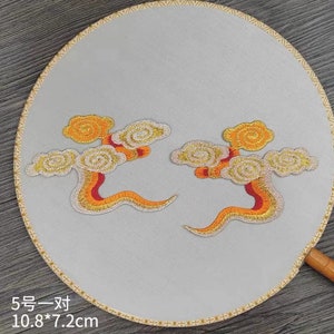 Può includere: Due toppe ricamate in oro e arancio con nuvole vorticose e accenti rossi. Le toppe misurano 10,8 x 7,2 cm e sono su uno sfondo bianco.