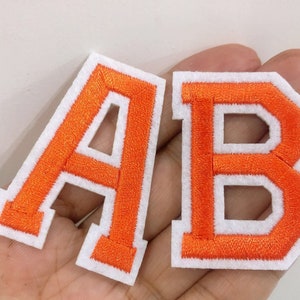 Könnte beinhalten: Zwei Filzpatches mit den Buchstaben A und B in Orange mit weißen Umrandungen. Die Buchstaben sind auf einem weißen Hintergrund aufgenäht.