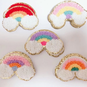 May include: Six rainbow-coloured felt patches with gold glitter trim. Each patch features a rainbow with fluffy white clouds. The rainbows are in different colour combinations, including red, orange, yellow, green, blue, purple, and pink.