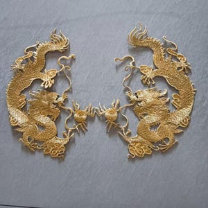 May include: Two gold embroidered dragon appliques. Each dragon is intricately detailed with scales, claws, and flowing manes. The dragons face each other, connected by a decorative element. The appliques are set against a grey background.
