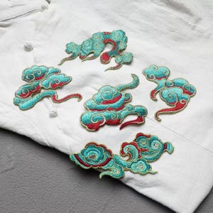 Peut inclure: Écussons de nuages brodés en fil turquoise, rouge et or. Les écussons sont de formes et de tailles variées, avec des détails complexes. Ils sont placés sur un fond de tissu blanc.