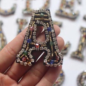 Könnte beinhalten: Ein schwarzer Filzbuchstabe A, der mit bunten Strasssteinen und Perlen besetzt ist. Der Buchstabe wird in einer Hand gehalten.