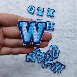 May include: A collection of embroidered letter patches featuring a gradient design that transitions from light blue to white, outlined with a dark blue border. The letters include Q, E, K, W, H, U, N, Z, and M, showcasing a chevron-like stitch pattern.