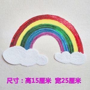 Puede incluir: Un parche de lentejuelas con forma de arcoíris con nubes blancas a cada lado. El arcoíris está hecho de lentejuelas rojas, naranjas, amarillas, verdes, azules, índigo y violetas. El parche mide 15 cm de alto y 25 cm de ancho.