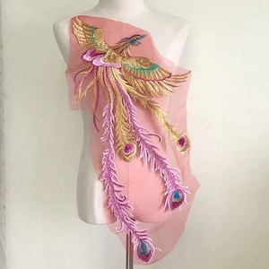 May include: A gold and pink embroidered phoenix applique with iridescent details. The phoenix is on a sheer pink fabric.