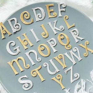 Può includere: Un set di lettere dell'alfabeto in oro e bianco su uno sfondo blu chiaro. Le lettere sono ricamate e hanno un carattere corsivo decorativo.