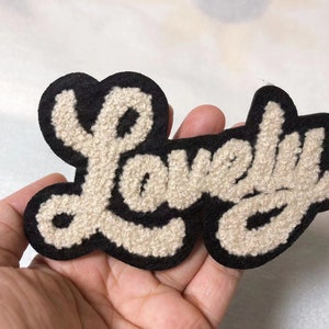 Pode incluir: Um remendo de feltro preto e branco com a palavra "Lovely" em uma fonte cursiva. As letras são em relevo e têm uma textura felpuda.