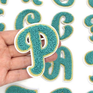 Puede incluir: Un conjunto de letras de fieltro color turquesa con contorno dorado. Las letras están en una fuente cursiva y son perfectas para agregar un toque de personalidad a cualquier proyecto.