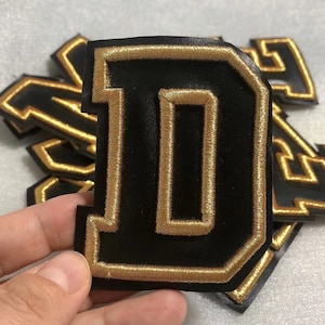 Op de afbeelding: Een zwarte viltletter D met gouden geborduurde stiksels. De letter is een patch en wordt vastgehouden door een hand.