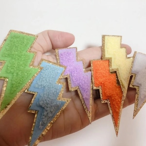 Può includere: Sei toppe a forma di fulmine con bordino dorato glitterato. Le toppe sono realizzate in tessuto peloso e sono nei colori verde, azzurro, viola, arancione, giallo e lavanda.