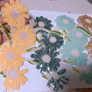 Op de afbeelding: Assortiment van kleurrijke daisy-vormige stoffen applicaties met pailletten. De applicaties zijn in tinten geel, groen, blauw en bruin.