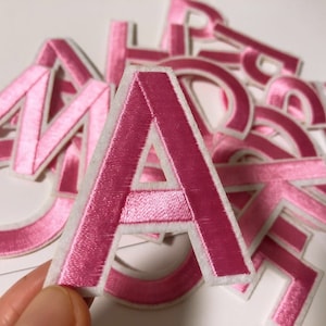 Pode incluir: Letras de feltro rosa com contornos brancos. As letras são A, M e C. As letras estão dispostas em uma pilha.