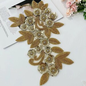 Pode incluir: Aplique floral dourado com contas e lantejoulas. O design apresenta folhas e flores douradas com lantejoulas prateadas e centros de strass transparentes. O aplique é provavelmente para embelezar roupas ou acessórios.