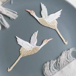 May include: Two embroidered crane patches in flight. The patches feature white wings, tan bodies, and pink beaks. The patches are on a gray plate with a white fringed cloth.