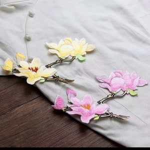 May include: Three embroidered fabric patches featuring magnolia flowers. The patches are in shades of yellow, pink, and white. The flowers are detailed with delicate stitching and have green leaves.
