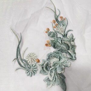 May include: Green and white embroidered floral applique featuring a large green rose and smaller white flowers. The applique is on a white background.