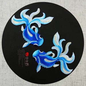 May include: Two blue embroidered fish with white accents on a black background. The fish are facing each other and have a traditional Chinese design.