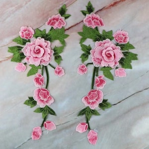 May include: Two pink floral appliques with green leaves and stems. The appliques are made of fabric and are embroidered with pink flowers and green leaves.