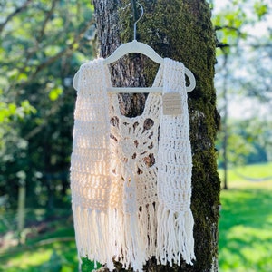 May include: A white crocheted vest with fringe hanging from the bottom. The vest is hanging on a white hanger against a tree trunk.