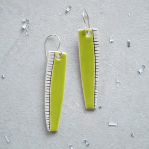 May include: A pair of lime green and white ruler-shaped earrings with silver hooks. The earrings have a bright green centre with black ruler markings on a white background. The earrings are displayed on a light grey surface with silver confetti.