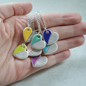 May include: A set of five ceramic heart necklaces with silver chains. Each heart is painted with a different colour and black stripes. The colours include yellow, green, pink, purple, and blue.