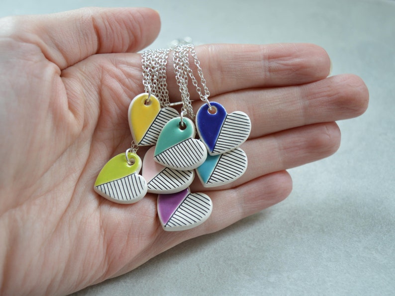 May include: A set of five ceramic heart necklaces with silver chains. Each heart is painted with a different colour and black stripes. The colours include yellow, green, pink, purple, and blue.