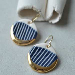 May include: A pair of round ceramic drop earrings featuring a white base with dark blue vertical stripes and a polished gold-tone metallic edge. Each earring hangs from a thin gold-tone wire hook, showcasing a smooth, glossy finish throughout.