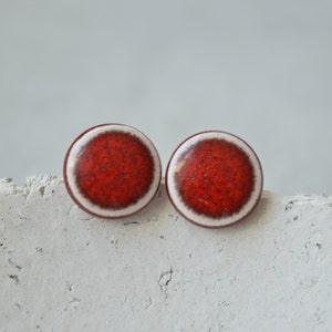 Op de afbeelding: Een paar rode en witte keramische oorbel studs. De oorbellen zijn rond en hebben een witte rand.