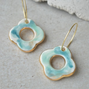 May include: A pair of gold hoop earrings with a light blue and white ceramic flower charm. The flower charm has a gold rim and a swirl pattern.