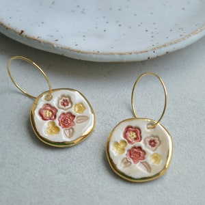 Könnte beinhalten: Ein Paar handgefertigte Keramikohrringe. Jeder Ohrring hat eine weiße, kreisförmige Basis mit geprägten Blumendesigns in Rot, Rosa und Gelb, die mit Gold umrandet sind. Die Ohrringe hängen an goldfarbenen Reifen.